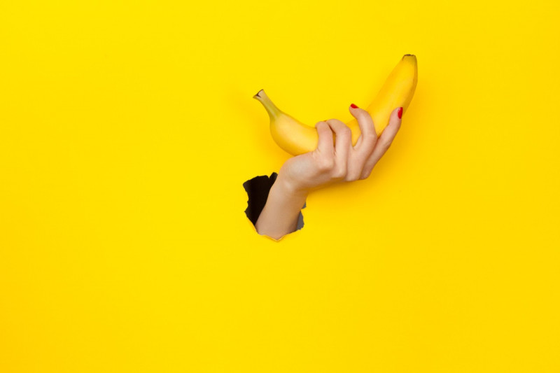 Hand holding a banana through a hole in a yellow background